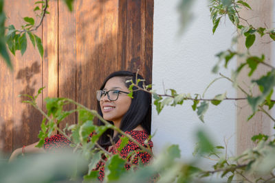 Thoughtful woman smiling while sitting against wall seen through leaves