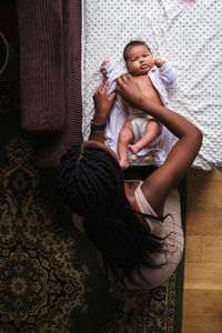 Top view of unrecognizable caring african american mother with long braided hair dressing bodysuit on newborn baby lying on blanket on sofa