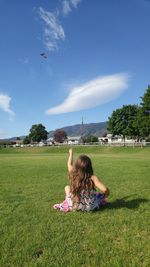 Rear view of girl flying kite while sitting on grassy field