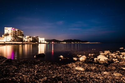 Illuminated buildings by sea against sky at night