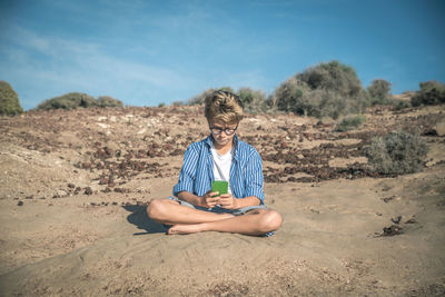 Boy using phone while sitting on field against blue sky