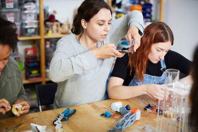 Young female inventor photographing model on table while sitting amidst colleagues at workshop