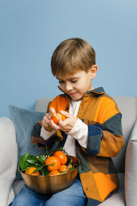 Boy cleans and eats a tangerine while sitting in a chair on a blue background.