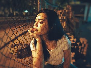 Thoughtful woman looking through chainlink fence while sitting outdoors at night