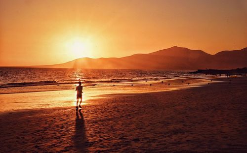 Rear view full length of man running at beach against orange sky