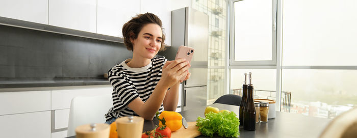 Portrait of young woman using mobile phone while sitting in kitchen