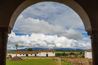 Cross sculpture and houses against cloudy sky seen through arch