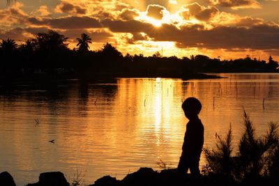 Silhouette boy standing by lake against sky during sunset