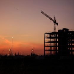 Silhouette crane by building against sky during sunset