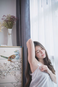 Mid adult woman with arms raised sitting against window at home