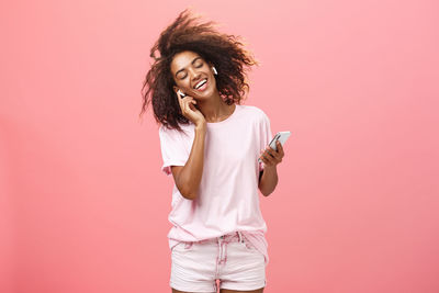 Beautiful young woman using phone while standing against pink background