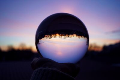 Close-up of hand holding crystal ball against sky during sunset