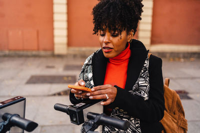 Young african american ethnic woman with curly hair in warm coat standing on city street and doing contactless payment for electric scooter with smartphone using application