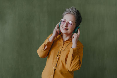 Smiling woman with eyes closed listening to music through headphones in front of green wall