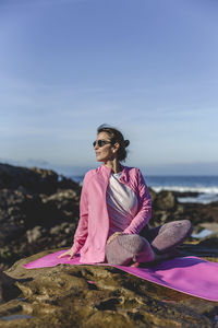 Mature woman exercising while sitting over mat on rock by sea against sky