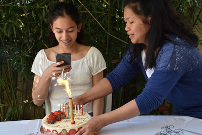 Smiling woman using mobile phone while woman holding birthday candle on table