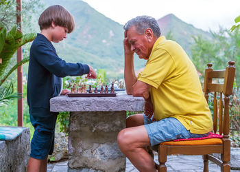 The boy is playing chess on the stone table with his grandfather in the backyard. 