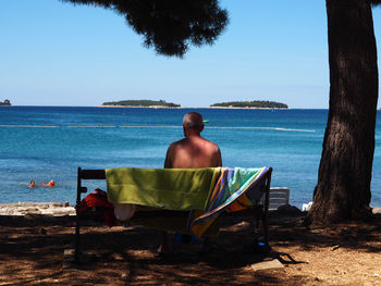 Rear view of shirtless man sitting on chair at beach