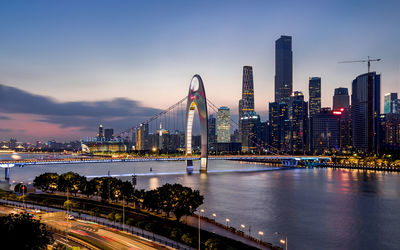 Illuminated bridge over river by city buildings against sky