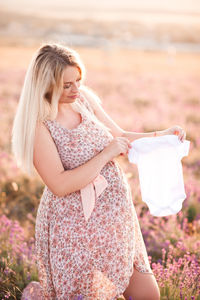 Smiling pregnant woman holding baby cloths standing on field