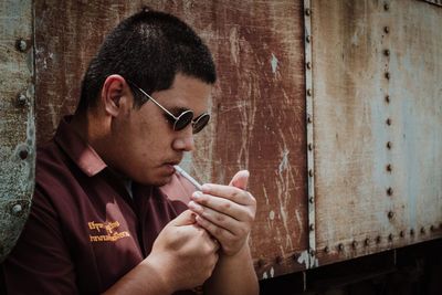 Man in sunglasses igniting cigarette against rusty wall