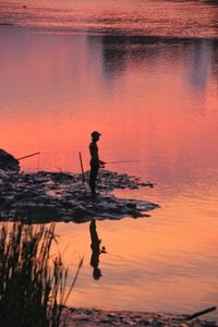 Silhouette man fishing while standing in lake against orange sky