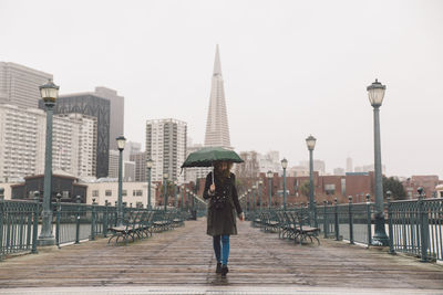 Full length of young woman holding umbrella walking on pier against transamerica pyramid