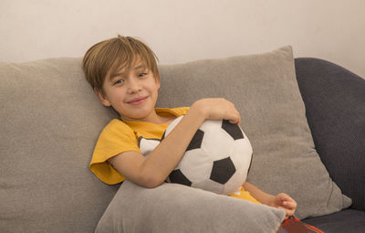 Portrait of boy holding soccer ball while sitting on sofa at home