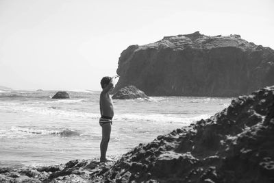 Side view of shirtless boy looking away while standing at beach on sunny day