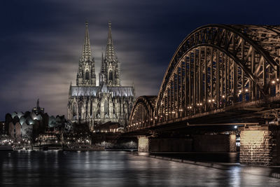 Illuminated bridge over river by buildings against sky at night