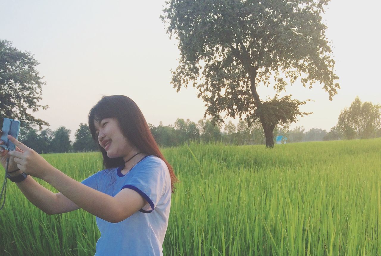 grass, lifestyles, leisure activity, tree, person, casual clothing, field, young adult, grassy, clear sky, smiling, three quarter length, childhood, growth, looking at camera, nature, landscape, green color