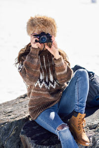 Young woman wearing warm clothing while photographing on rock during winter