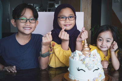 Portrait of smiling siblings with cake on table gesturing heart shape