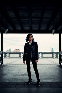 Full length portrait of young woman standing against bridge