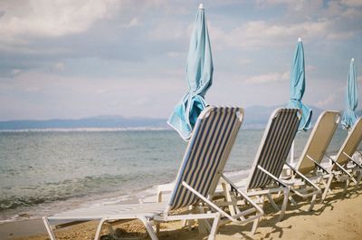 Deck chairs and closed parasols at beach against sky