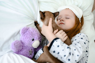 A little girl with red hair sleeps on a bed with white sheets and hugs her ginger cat in a dream. 