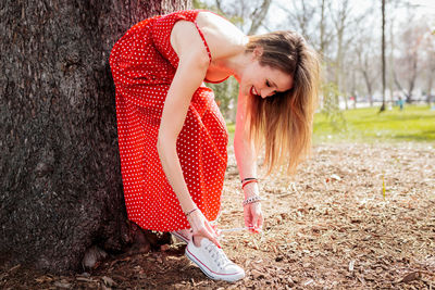 Smiling woman in red dress tying shoelace against tree trunk