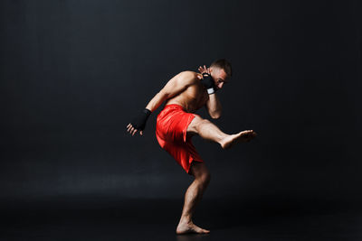 Full length of shirtless man kickboxing against black background