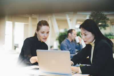 Confident businesswoman discussing over laptop with female colleague at table during meeting in cafeteria