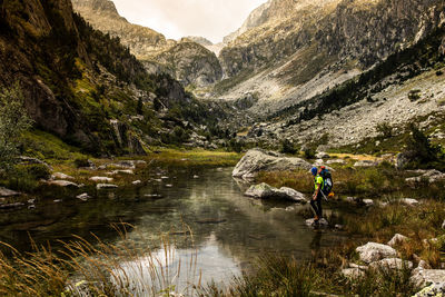 Side view of unrecognizable adventurer with backpack standing on stone amidst shallow river flowing through rocky pyrenees mountains in summer day