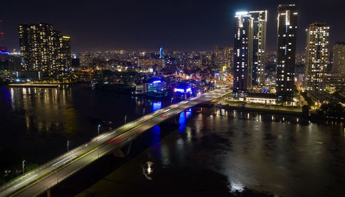 Illuminated bridge over river by buildings against sky at night