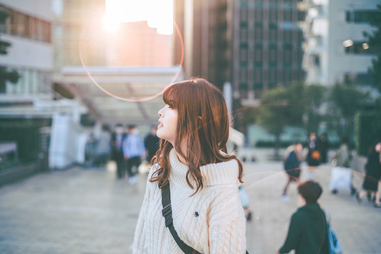 city, architecture, building exterior, real people, women, focus on foreground, lifestyles, built structure, one person, adult, street, incidental people, standing, leisure activity, city life, young women, sunlight, young adult, day, hairstyle, hair, outdoors, lens flare, beautiful woman, warm clothing