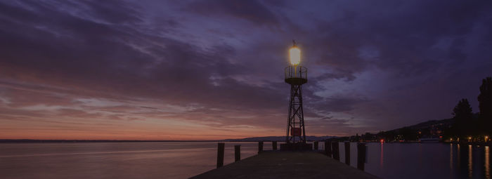 Illuminated tower by sea against sky at sunset