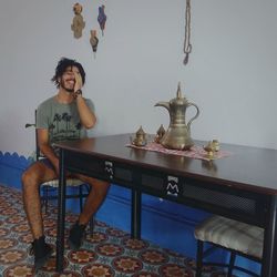 Smiling young man sitting by table against wall at home
