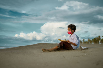 Boy wearing mask writing in paper while sitting at beach against sky