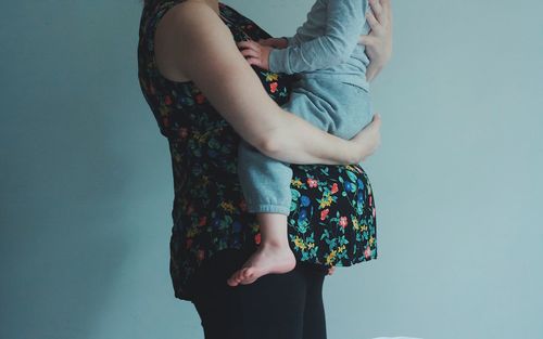 Cropped image of pregnant woman carrying son while standing against wall