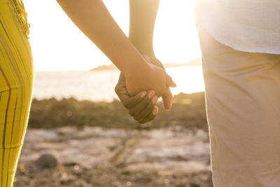 Midsection of couple holding hands while standing at beach during sunset