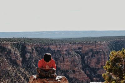Boy mediating while sitting on rock formation against clear sky