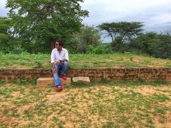 Young man sitting on retaining wall against trees