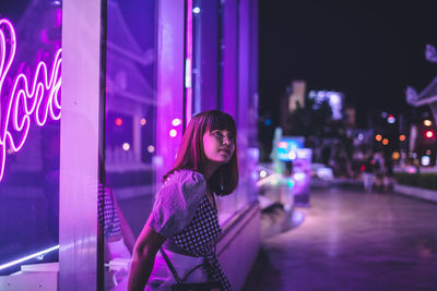 Young woman standing by illuminated building in city at night